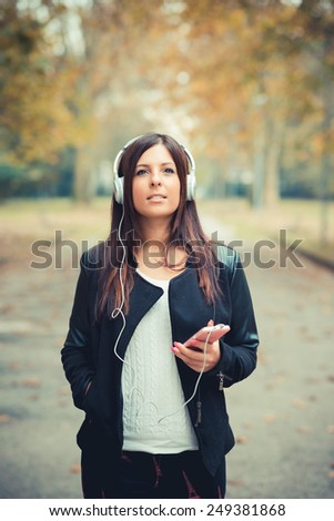 young beautiful brunette straight hair woman in the park during autumn season - listening to music with headphones and smartphone