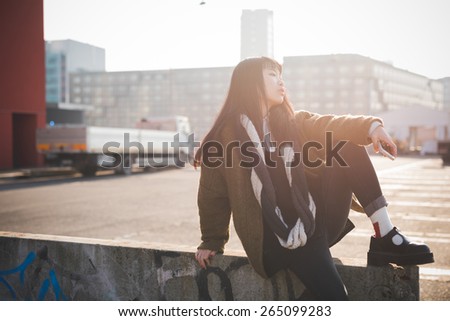 young beautiful asian hipster woman in the city - living town during sunset in backlit - sitting on a wall with smartphone - concept emancipation and future