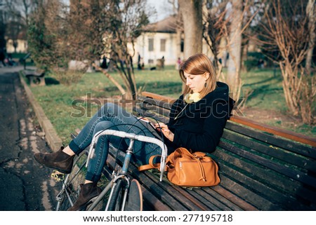 young beautiful hipster sporty blonde woman in town with bike using smartphone sitting in a bench
