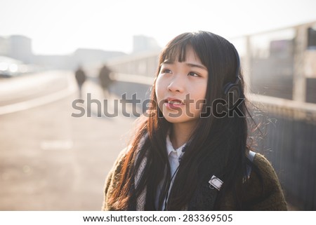 young beautiful asian hipster woman in the city - living town during sunset in backlit - listening music with headphones