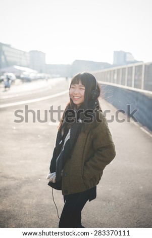 young beautiful asian hipster woman in the city - living town during sunset in backlit - listening music with headphones