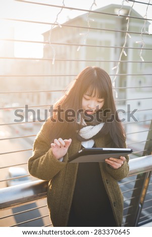 young beautiful asian hipster woman in the city - living town during sunset in backlit - using device wireless tablet online