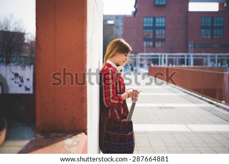 young beautiful blonde hipster woman in the city with ukulele using smartphone connected online