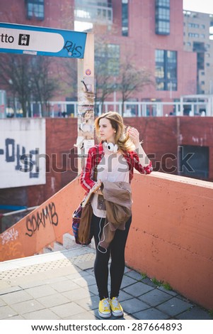 young beautiful blonde hipster woman in the city using smartphone connected online with headphones