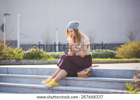young beautiful blonde hipster woman in the city with ukulele using smartphone connected online