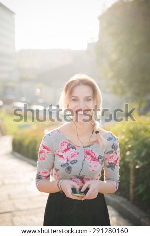 Young beautiful blonde caucasian girl walking through the city listening to music with earphones and smartphone, wearing blue and pink dress with black skirt - technology, music, relax concept