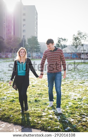 A couple of young lovers in the city, hand in hand. She is wearing black jeans and a blue shirt. He is wearing blue jeans and a red striped sweater.