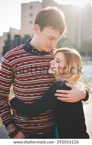 A couple of young lovers embraced in the city. She is wearing black jeans and a black sweater. He is wearing blue jeans and a red striped sweater.