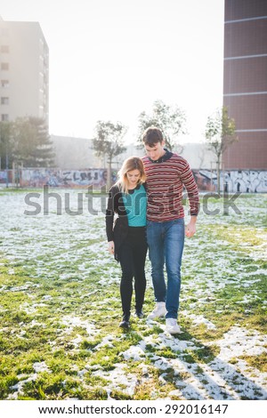 A couple of young lovers embraced in the city.  She is wearing black jeans and a blue shirt. He is wearing blue jeans and a red striped sweater.