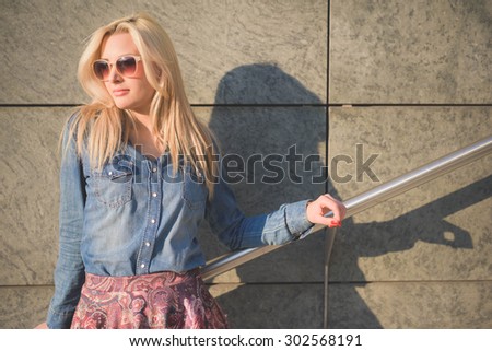 Half length of a young beautiful blonde caucasian girl posing leaning on a wall in the city wearing a jeans shirt and a floral skirt and a pair of sunglasses overlooking right