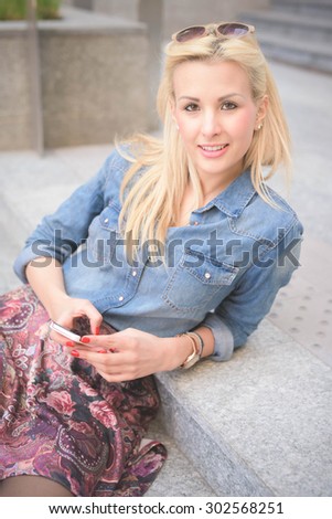Half length of a young beautiful blonde caucasian girl seated on a staircase using a smartphone looking in camera - communication, technology, social network concept
