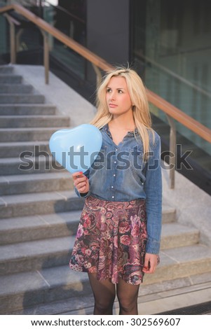 Knee figure of a young beautiful blonde girl posing leaning on a wall with a hearted balloon outdoor in the city overlooking on her right wearing a jeans shirt and a floral skirt