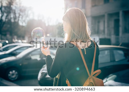 Half length view from back of young beautiful hipster sporty blonde woman in town playing with bubble soap - childhood, happiness, carefreeness concept - backlight
