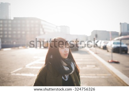 Half length of young beautiful asian long brown straight hair hipster woman, overlooking left, pensive, in the city during sunset in backlight- thoughtful, thinking future concept