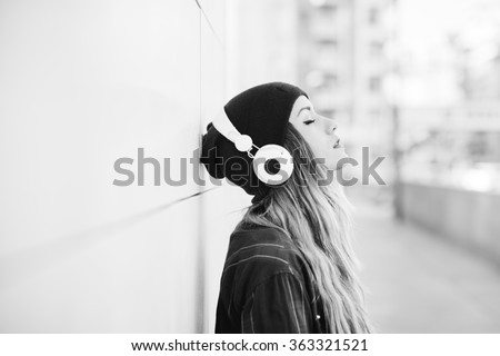 Black and white half length profile portrait of young handsome caucasian blonde hair woman leaning against a wall, listening music with headphones, eyes closed - serene, enjoying, music concept