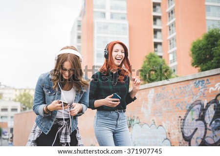 Knee figure of two young handsome caucasian blonde and redhead straight hair women dancing in the city listening music with headphones and smartphone, laughing - music, fun, freedom concept