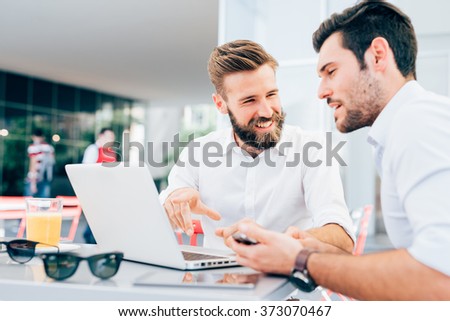 Two young bearded caucasian modern business man sitting in a bar, using laptop, looking each other, smiling and chatting - business, work, technology concept
