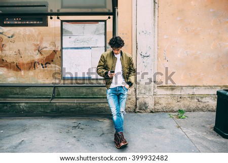 Young handsome caucasian man leaning on a bus stop holding a smart phone looking down and tapping the screen - technology, social network, communication concept