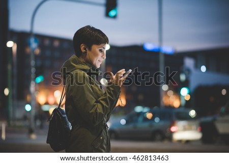 Half length of young handsome caucasian brown straight hair woman holding a smartphone looking down the screen in city night, face illuminated by screen light - technology, communication concept