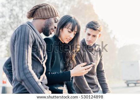 Three multiethnic woman and men friends outdoor in city back light, chatting and holding smart phone - friendship, social network, conversation concept