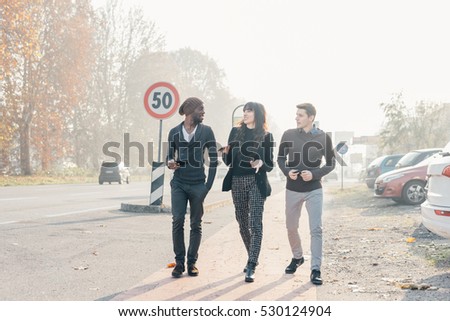 Three multiethnic woman and men friends walking outdoor in city back light, chatting and holding smart phone - friendship, social network, conversation concept