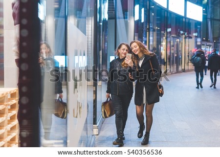 Two young beautiful caucasian women friends outdoor in the city night using smart phone hand hold strolling - technology, social network communication concept