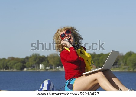 Beautiful woman, with laptop, laughs as she talks on the phone at the beach.