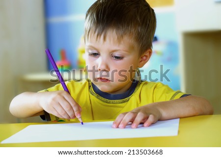 The boy draws a pencil at the table covered with a white tablecloth