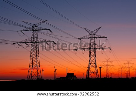 High voltage power lines with electricity pylons at twilight. At the horizon a nuclear power plant and wind turbines.