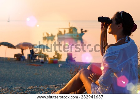 Girl with binoculars on the coast ; Young woman with binoculars watching a beautiful sunset on the beach of the Mediterranen Sea