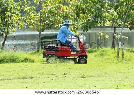 Workers on ride-on lawn mower cutting grass.