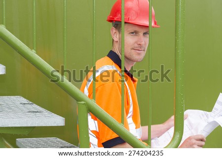 Portrait of confident architect holding blueprint while sitting by railing at storage tank park