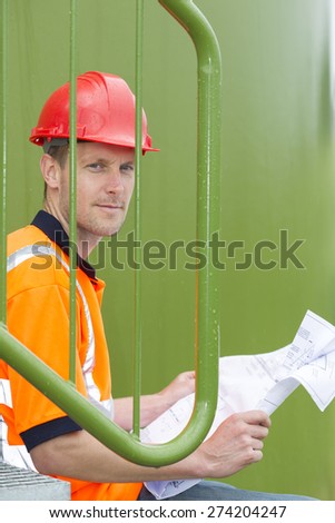 Portrait of mid adult architect holding blueprint while sitting in storage tank park