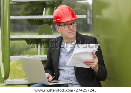 Mid adult architect holding blueprints while using laptop in storage tank park