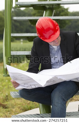Mid adult male architect in blazer analyzing blueprint while sitting on steps at storage tank park