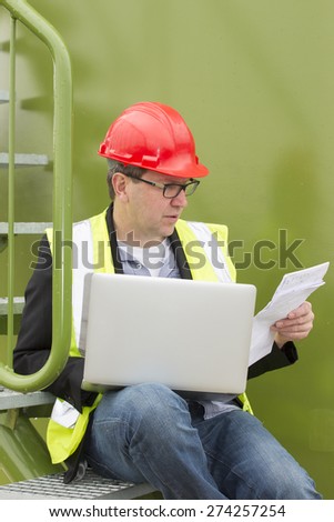 Mid adult architect holding blueprints while using laptop at storage tank park