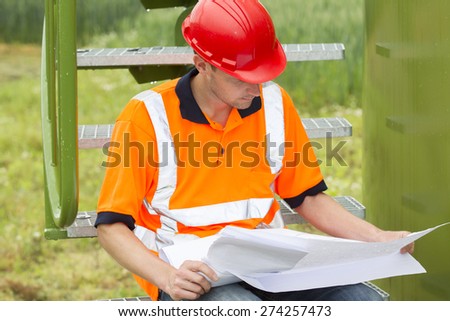 Mid adult male architect analyzing blueprint while sitting on steps in storage tank park