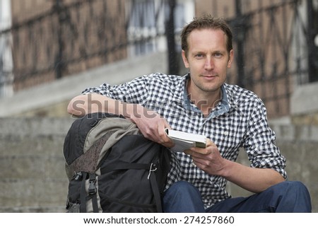Portrait of handsome mid adult tourist with book and backpack sitting against buildings in city