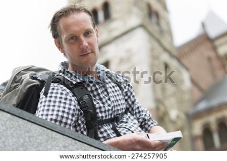 Low angle portrait of confident male tourist holding book while carrying backpack in city