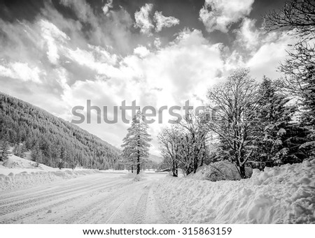 Beautiful landscape of a mountain scene with forest and road covered by snow; scattered cloudy sky; black and white conversion