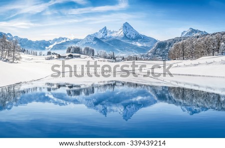 Panoramic view of beautiful white winter wonderland scenery in the Alps with snowy mountain summits reflecting in crystal clear mountain lake on a cold sunny day with blue sky and clouds