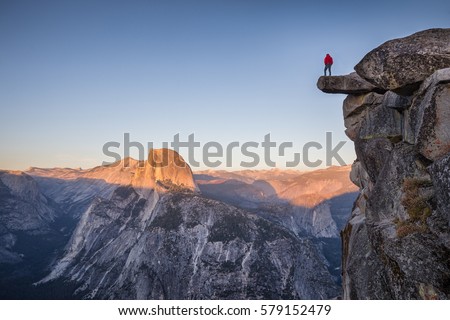 A fearless male hiker is standing on an overhanging rock at Glacier Point enjoying the view over Half Dome at sunset in beautiful twilight at sunset in summer, Yosemite National Park, California