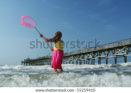 Beautiful young girl playing on the beach near the ocean. Fishing and looking for sea life using a cute pink net to match her dress. Rustic fishing pier in the background.
