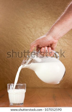 Hand pouring milk from jug to glass on brown background
