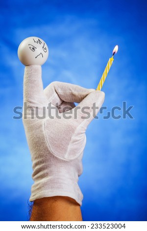 Burning birthday candle hold by angry white finger puppet over blue background