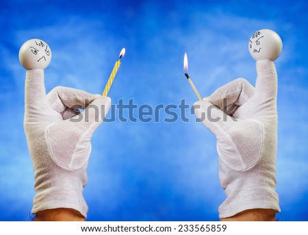 Burning birthday candle hold by angry white finger puppet against confused puppet with burning matchstick