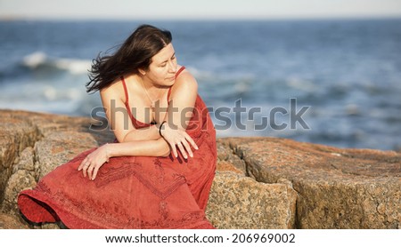 A beautiful woman in a red dress sits on a large rock at a beach during sunset.