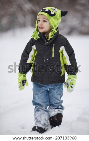 A young boy in a monster hat and gloves walks cheerfully through the snow.
