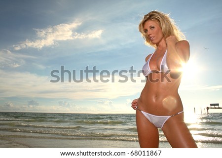 Blonde woman at the beach in her bathing suit. Looking  at the camera.Sun, water and sky in the background.