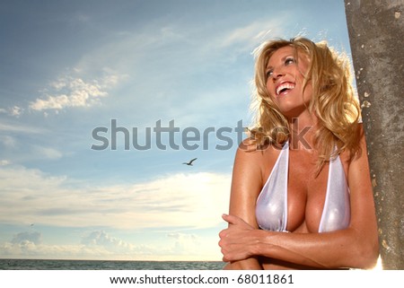 Blonde woman at the beach bikini. Laughing off camera. Blue sky and water in the background.
